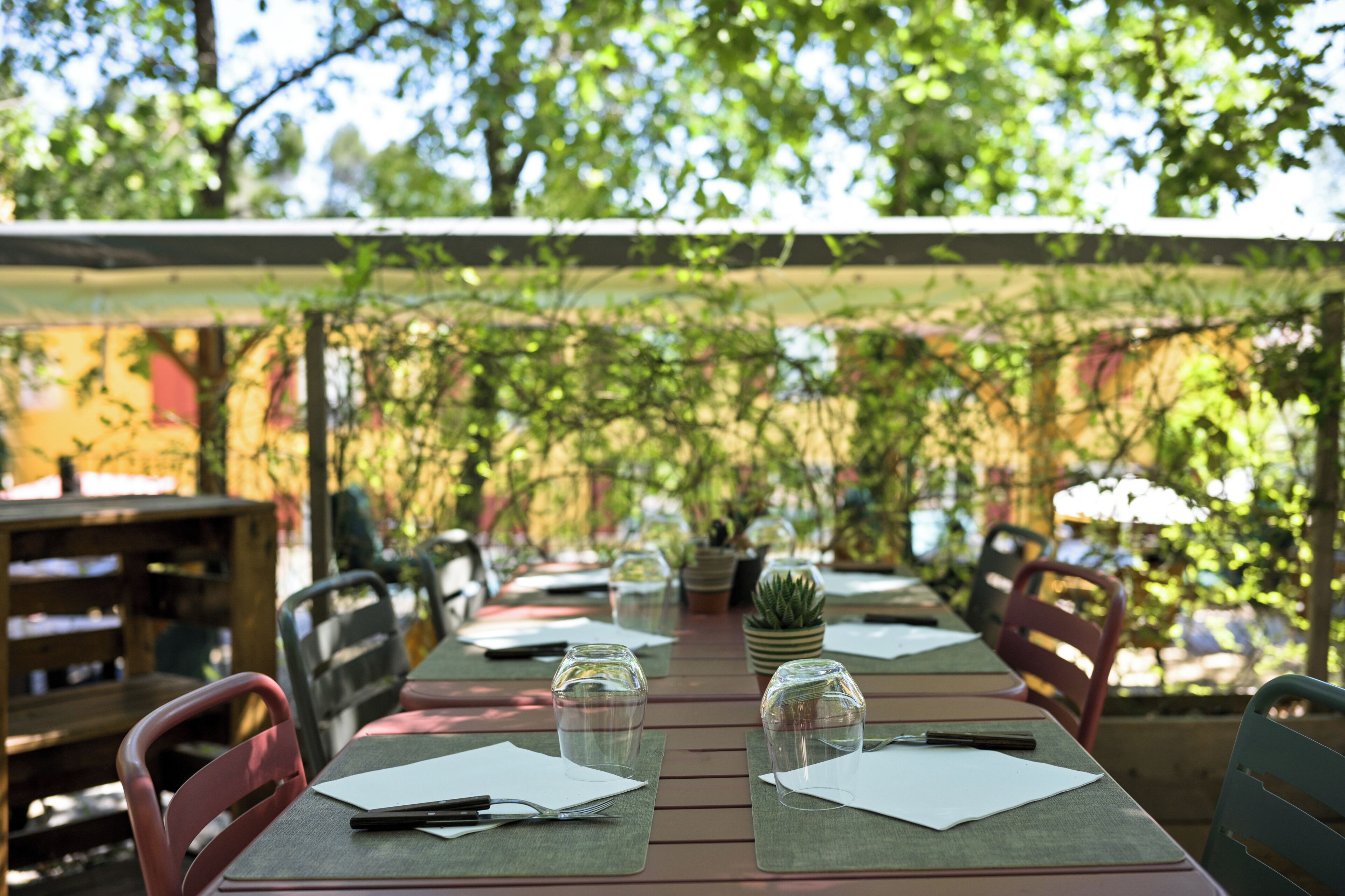 An outdoor dining area with tables set for two, each with a glass, napkin, and fork.