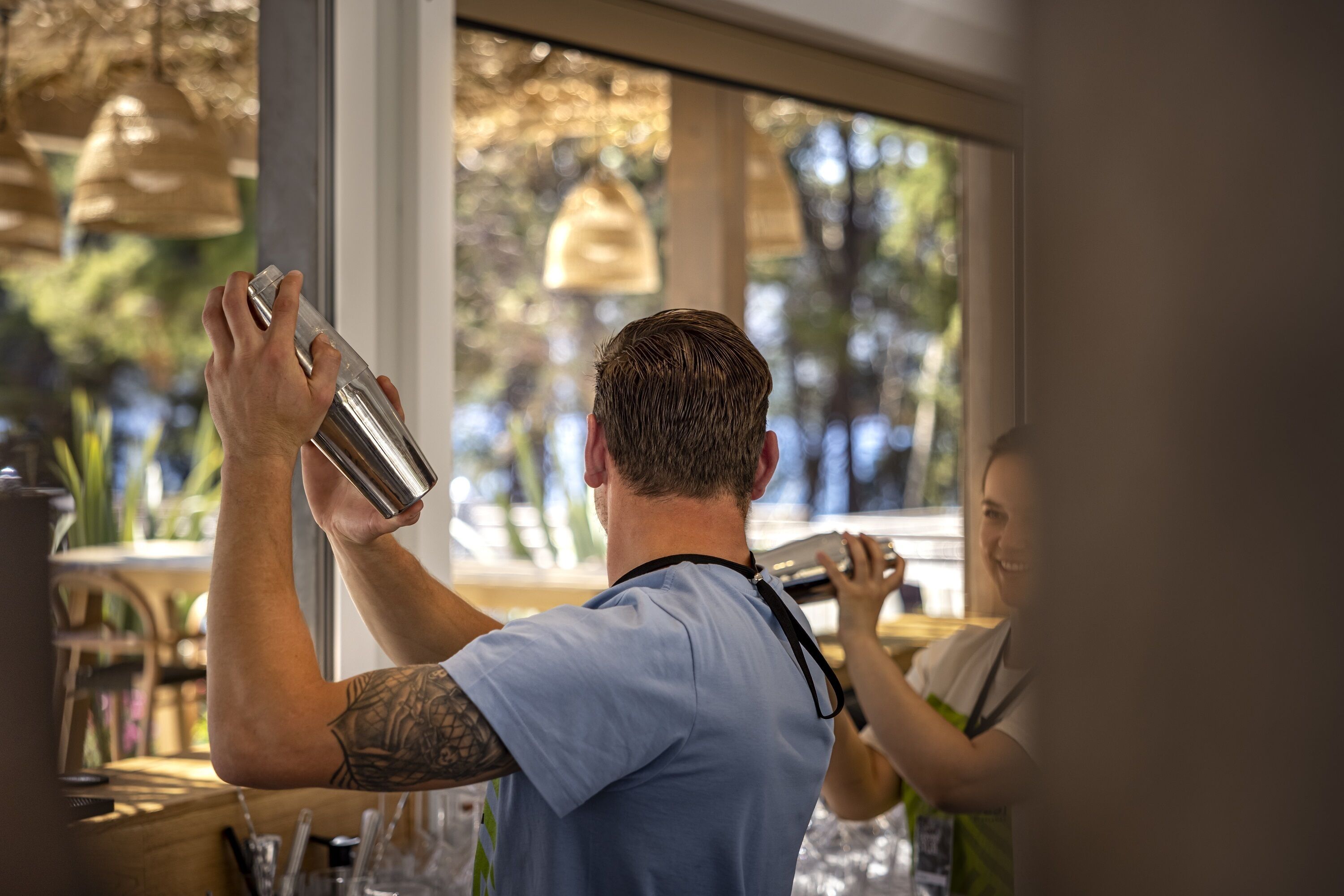 A person in a blue shirt preparing a drink behind a bar with a view of the outdoors through a window.