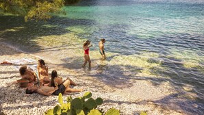 A group of people relaxing on a pebbled beach with clear water and greenery in the background.