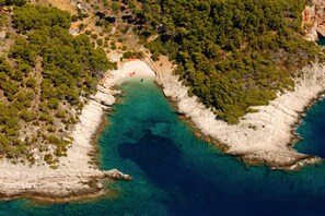 An aerial view of a coastal area with clear turquoise waters, rocky terrain, and dense green vegetation.