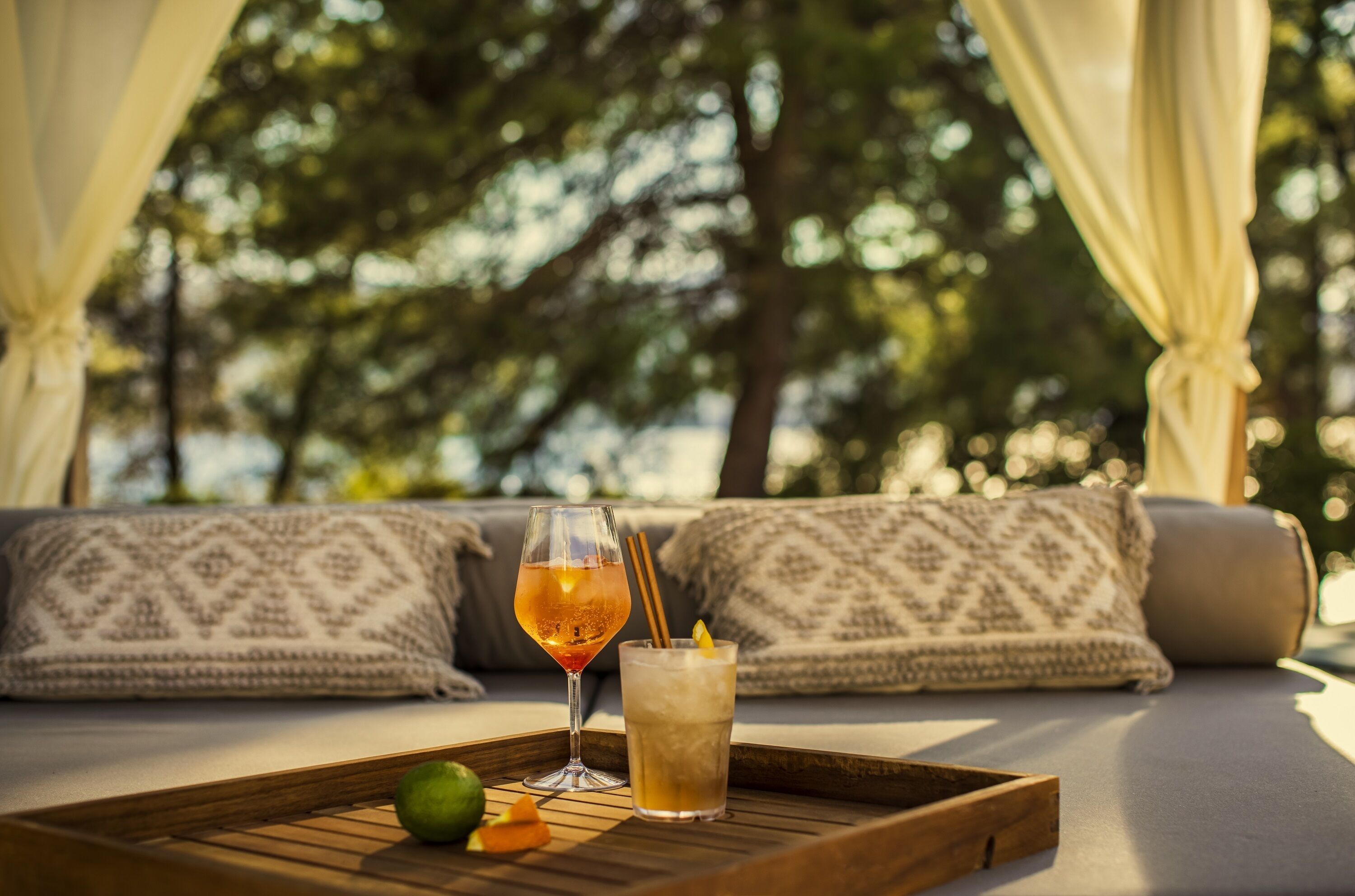 A tray with drinks and a lime on a wooden table in a cozy outdoor seating area with patterned cushions and a view of trees.