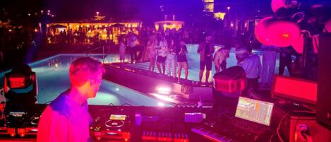 A DJ booth with electronic equipment and a poolside area with people and a bar in the background.