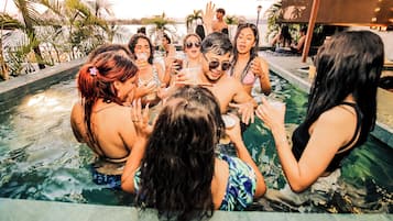 A group of people in a swimming pool with a tropical setting, including palm trees and a sunset in the background.