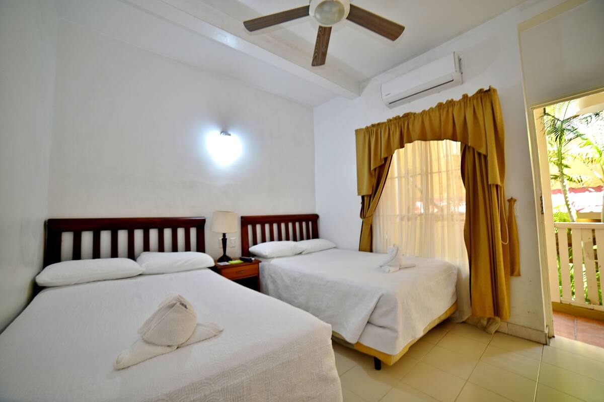 Habitación de hotel con dos camas individuales, ventilador de techo, aire acondicionado, mesita de noche con lámpara y vistas al exterior a través de una ventana con cortinas.