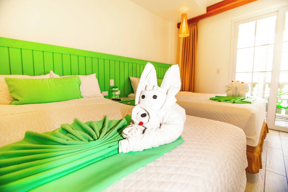 Habitación de hotel con dos camas, cabecera verde y una escultura de conejo hecha con toallas sobre la cama.