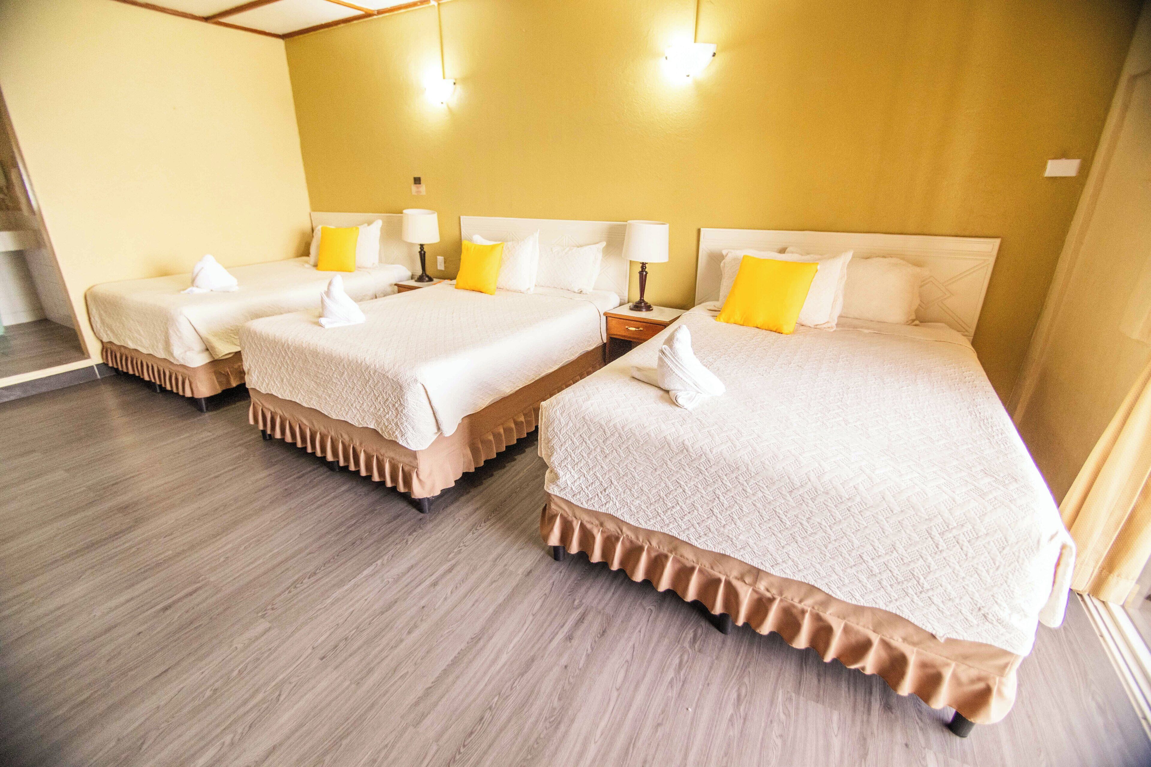 Habitación de hotel con tres camas, cada una con cubrecama blanco y falda con vuelo. Hay mesitas de noche con lámparas y un ventanal grande en un lateral.