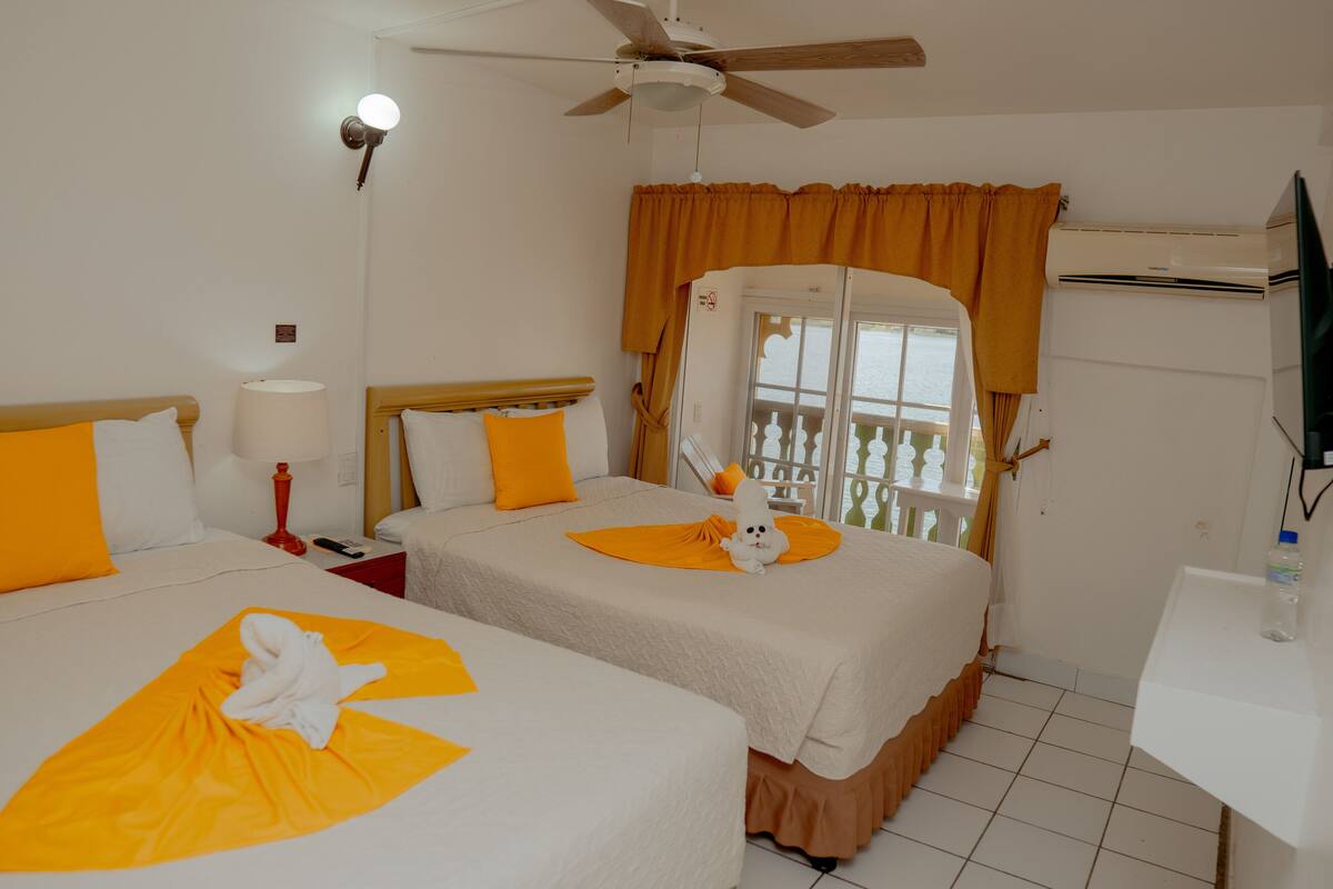 Habitación de hotel con dos camas, ventilador de techo, ventana con cortinas, televisor y una mesita.
