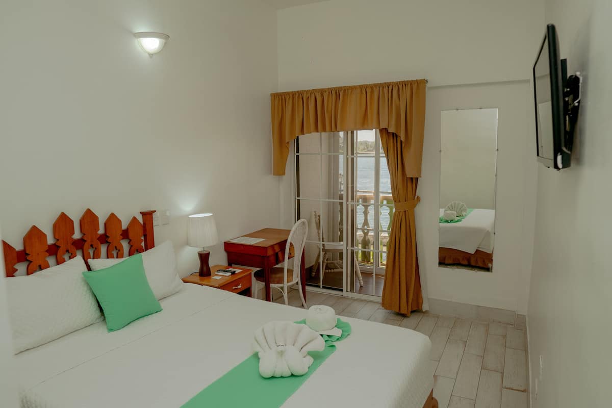 Habitación de hotel con cama, escritorio, silla, ventana con cortinas, vista al mar y televisor de pared.