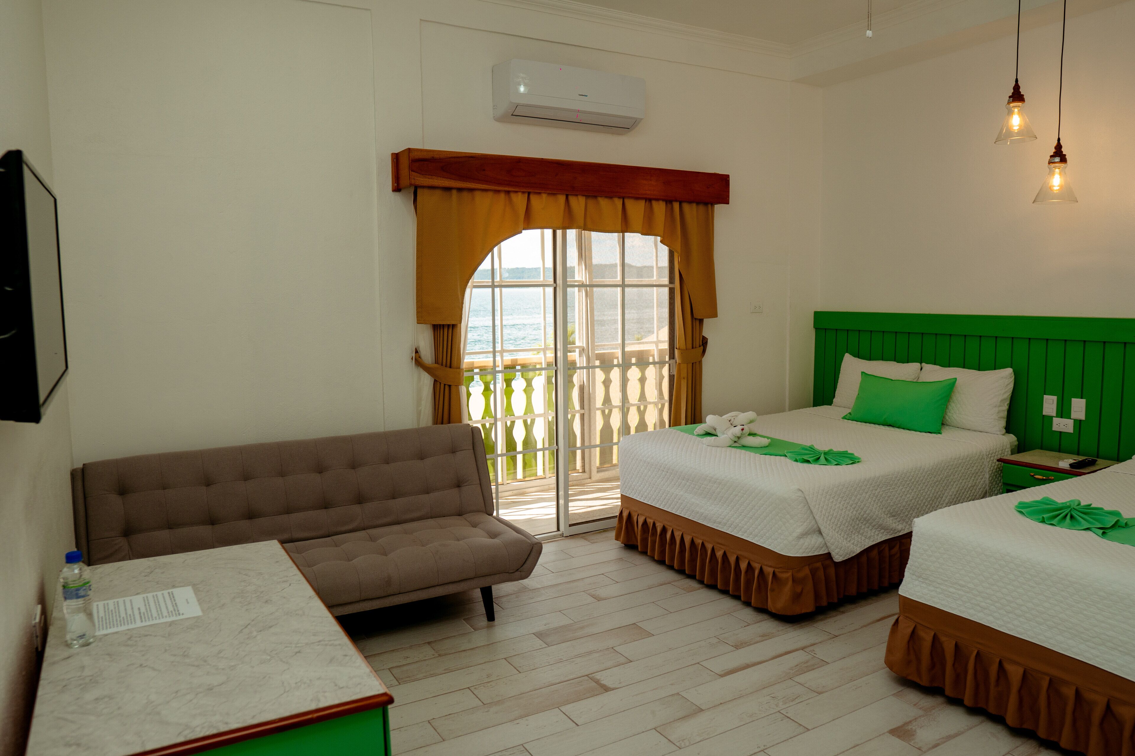A hotel room with a sofa, two beds, a window with curtains, a green headboard, and a marble table.