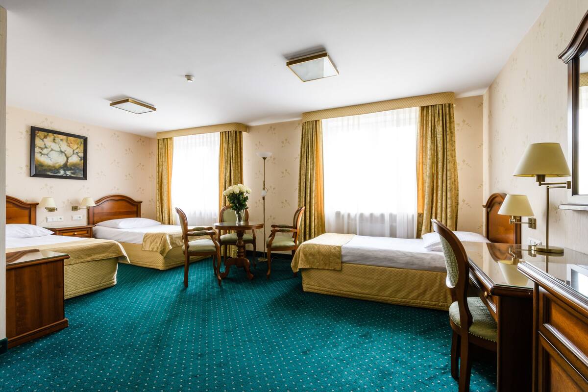 Habitación de hotel con dos camas individuales, un escritorio, una silla, una mesita con un florero y un cuadro en la pared.