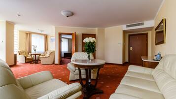 Et hotelværelse med rødt tæppe, beige sofaer, et rundt bord med en vase blomster og en dør, der fører til et andet rum.