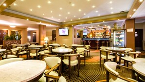 Un hall de hotel con mesas y sillas redondas, una zona de bar y una pantalla de televisión.