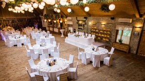 Un gran salón con múltiples mesas redondas preparadas para una recepción de boda, con manteles blancos y sillas, suelo de madera y una pared de ladrillo rústico con una exhibición de pan.