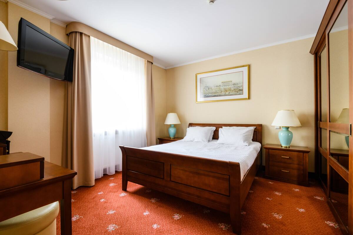 Habitación de hotel con cama, dos mesitas de noche con lámparas, televisión montada en la pared, escritorio y una silla.