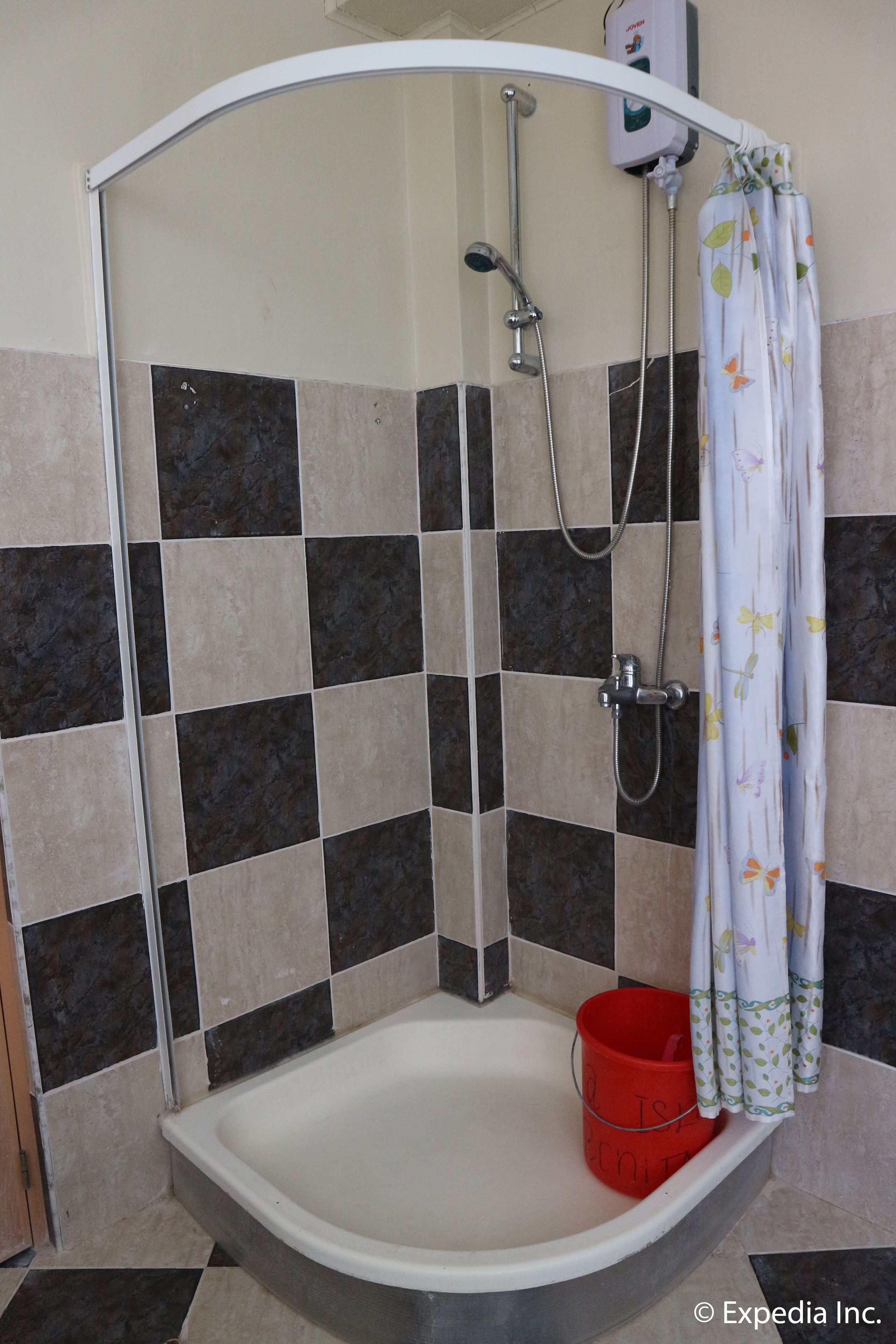 A shower area with a curved shower enclosure, a handheld showerhead, a wall-mounted faucet, a red bucket, and a shower curtain with a floral pattern.