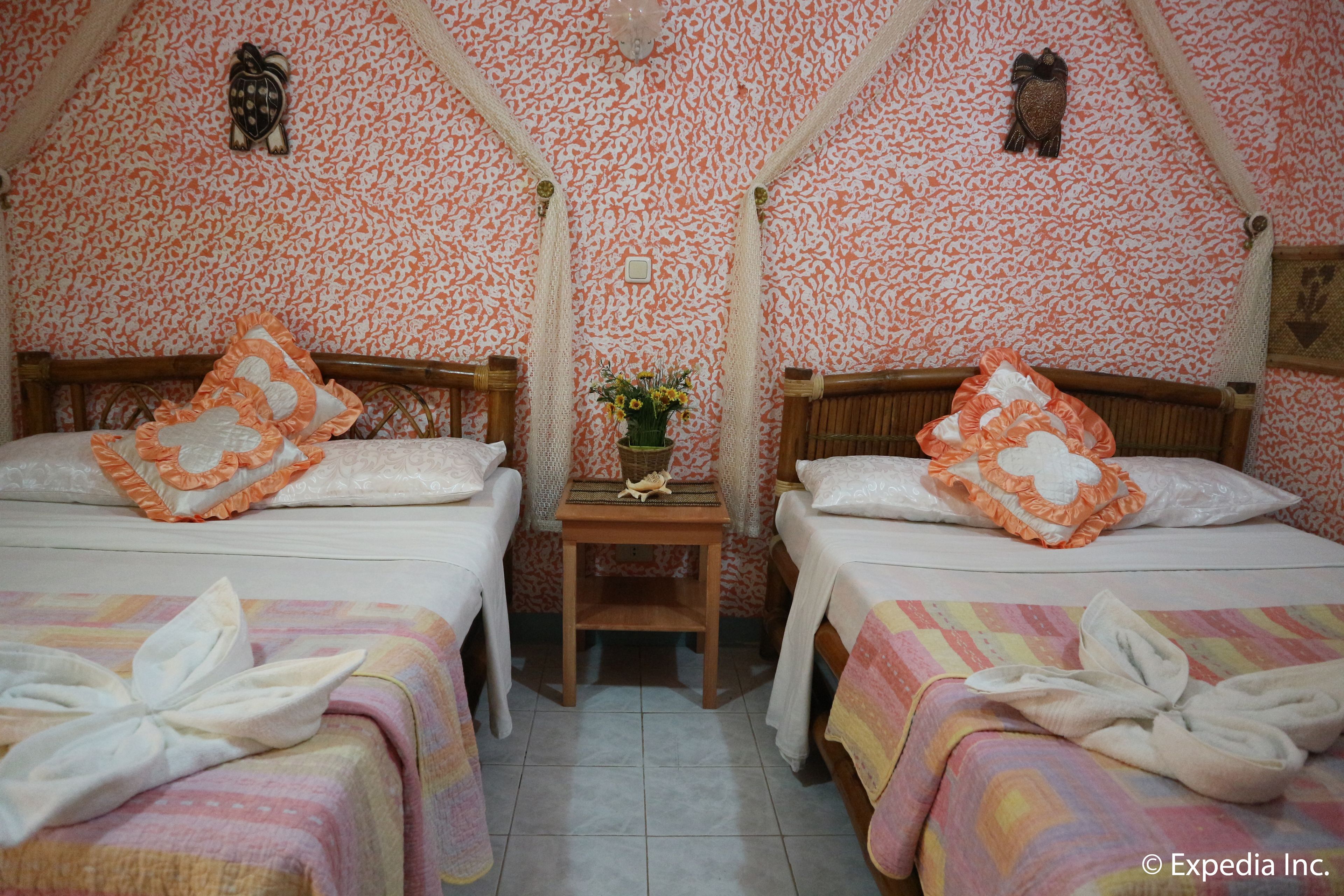Una habitación con dos camas, cada una con cabecero de madera, ropa de cama blanca y almohadas decorativas anaranjadas. Una pequeña mesa de madera con un jarrón de flores está entre las camas.