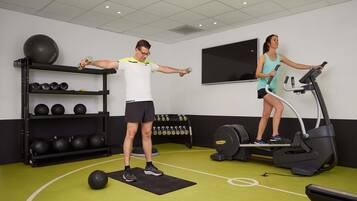 Een fitnessruimte met trainingsapparatuur, een tv aan de muur en een plank met fitnessballen.