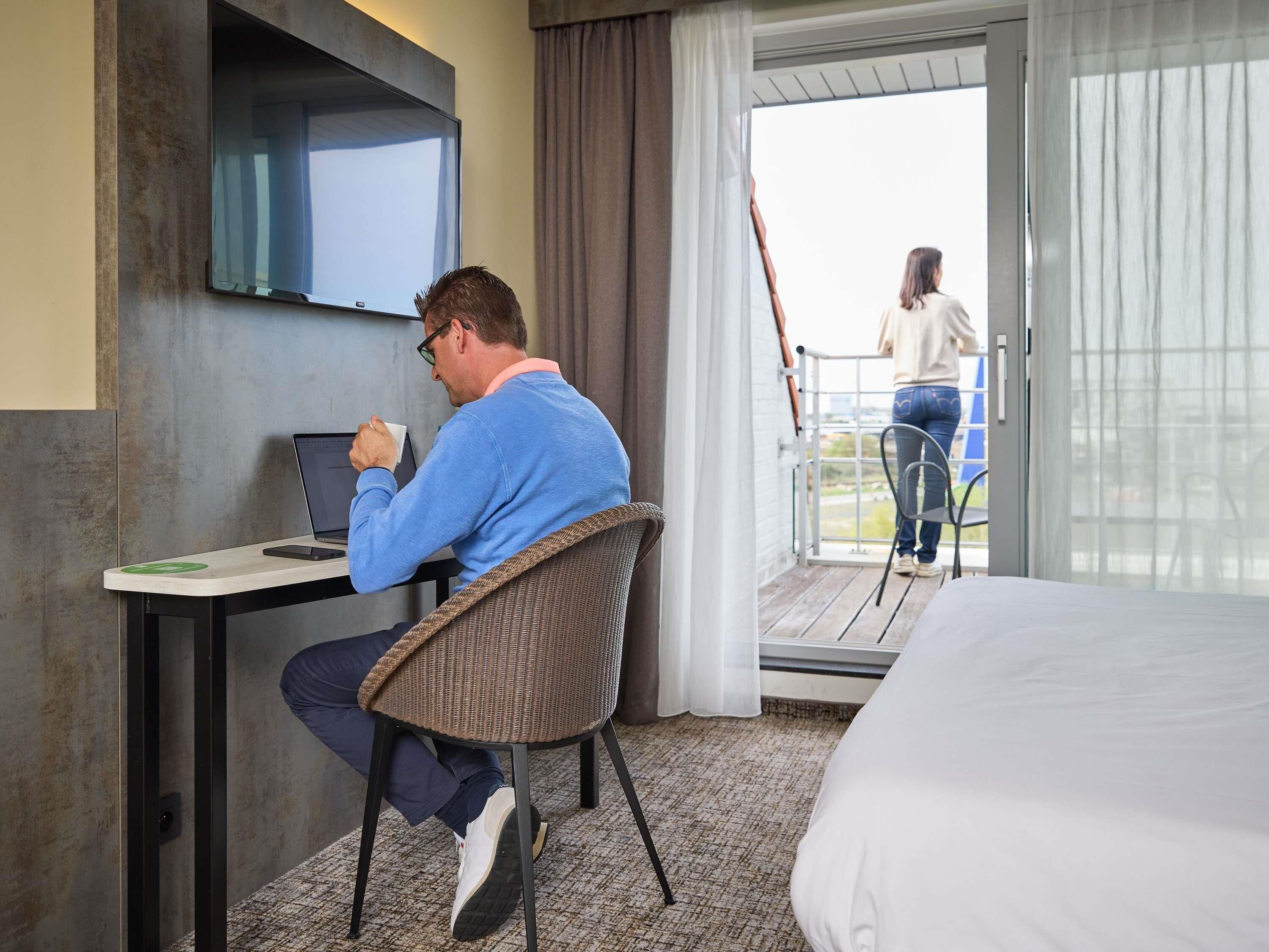 A hotel room with a bed, a desk with a laptop, a chair, a TV mounted on the wall, and a balcony with a railing.