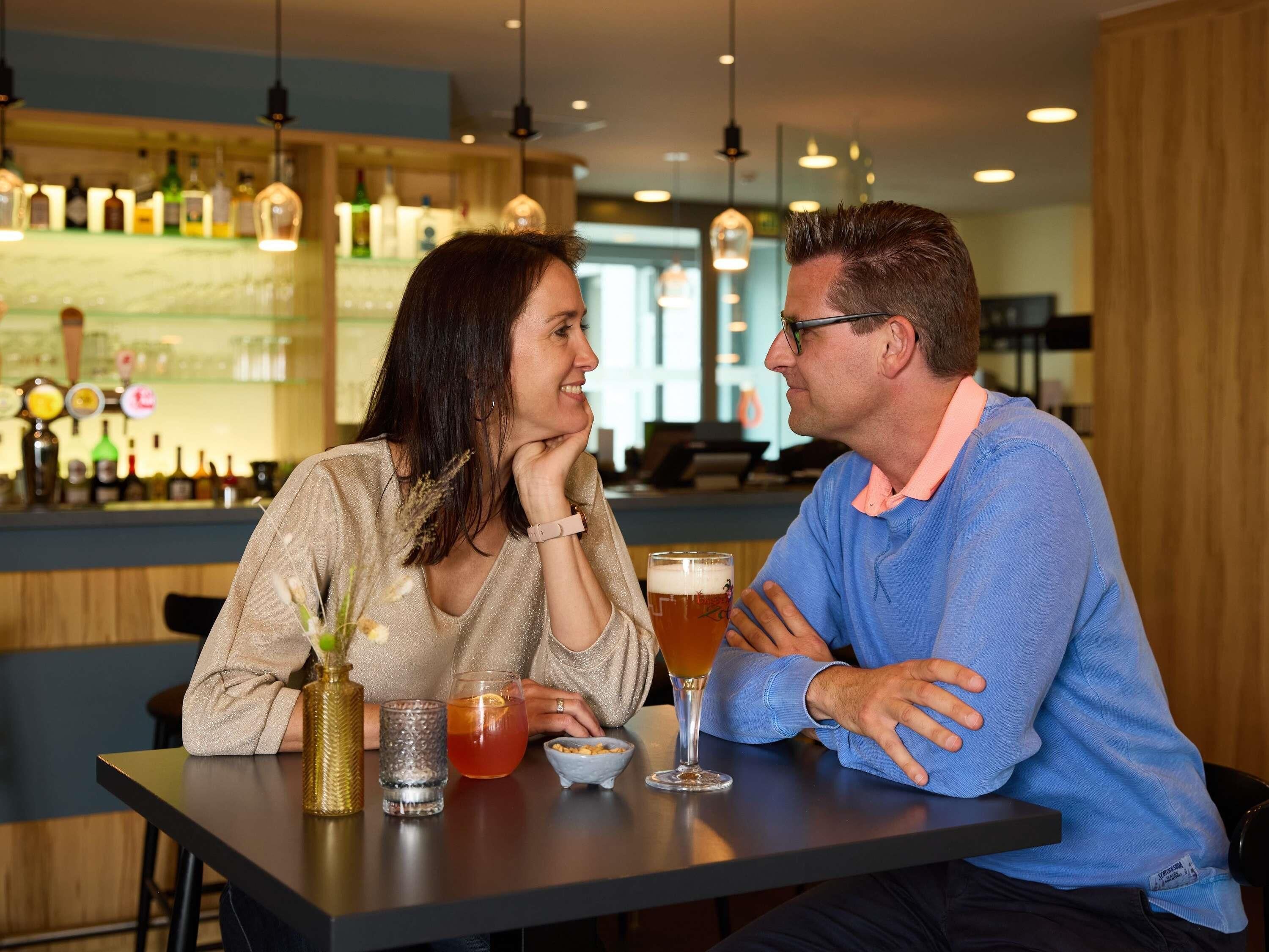 Dos personas sentadas en una mesa con bebidas, y un bar al fondo.