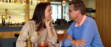 Dos personas sentadas en una mesa con bebidas, y un bar al fondo.