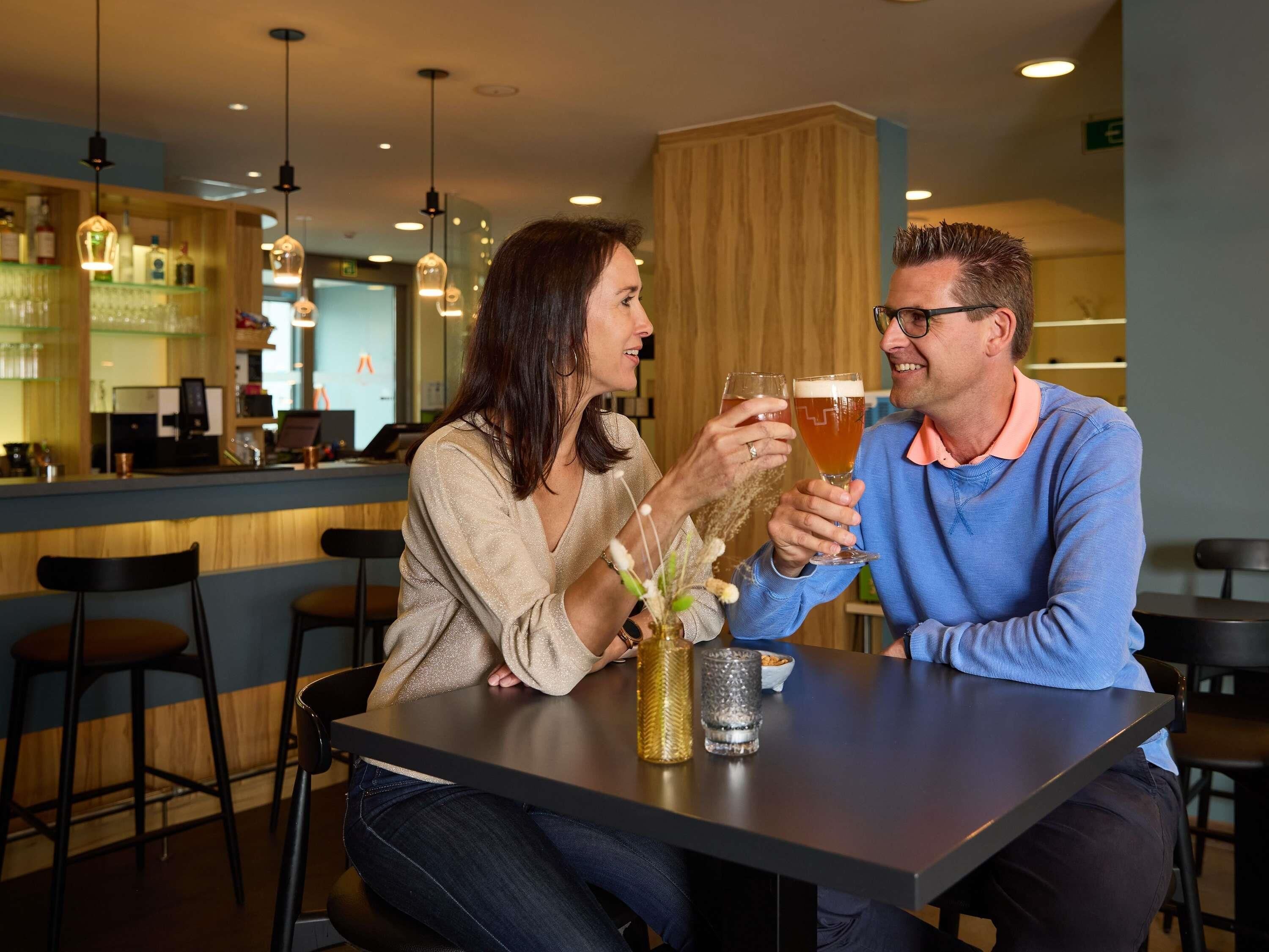 Twee personen zitten aan een tafel in een restaurant, met hangende lampen en een bar op de achtergrond.