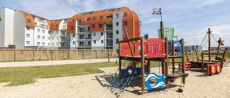 A playground with a swing set and climbing structures, adjacent to a building with orange roofs and multiple balconies.