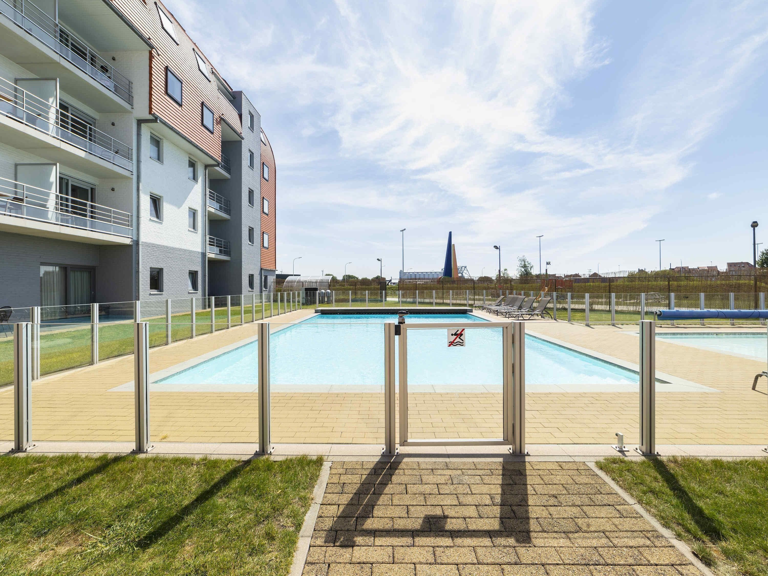 Una piscina con valla, zona verde y un edificio de varios pisos al fondo.