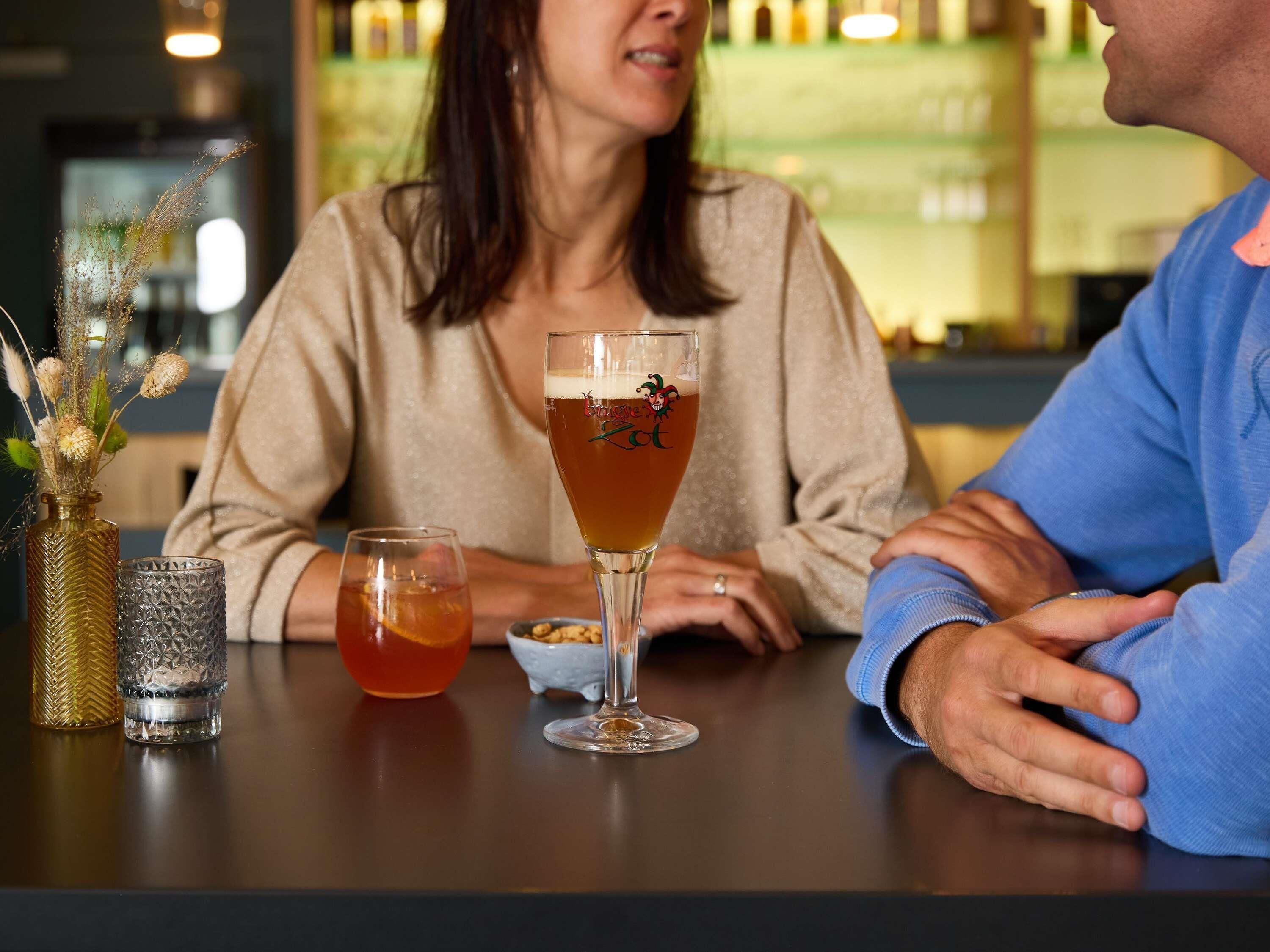 Un bar con una copa de bebida ámbar, un jarrón decorativo con flores secas y un vaso texturizado.