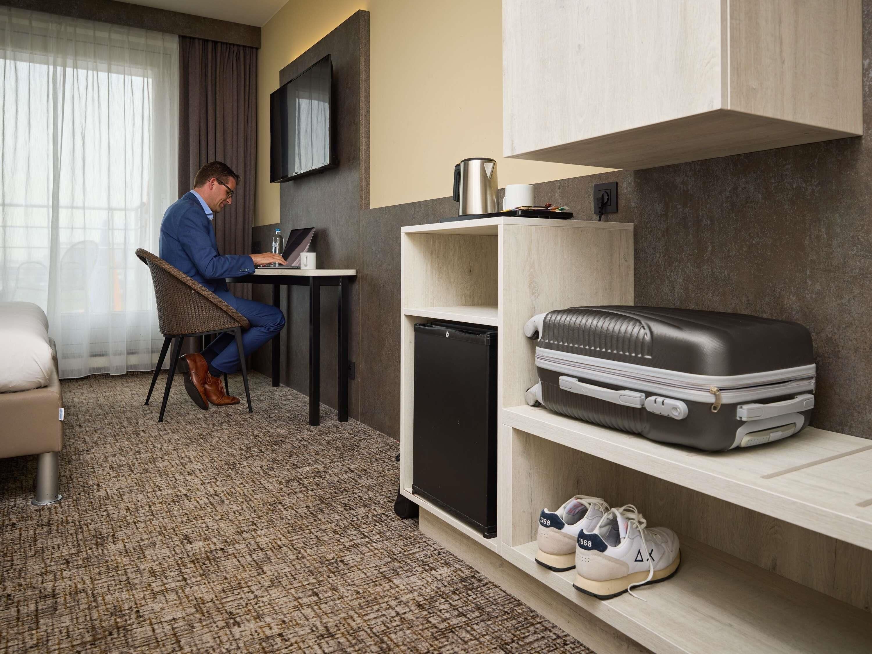 A hotel room with a desk, chair, television, minibar, and a suitcase on a shelf.