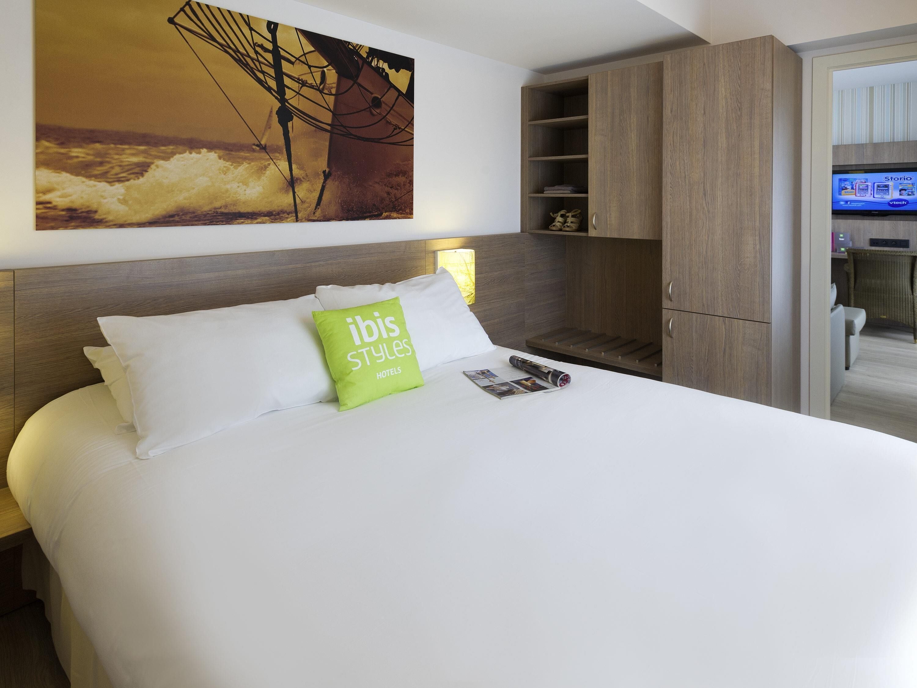 A hotel room with a bed, a green pillow, a wooden headboard, a TV, and a wardrobe.
