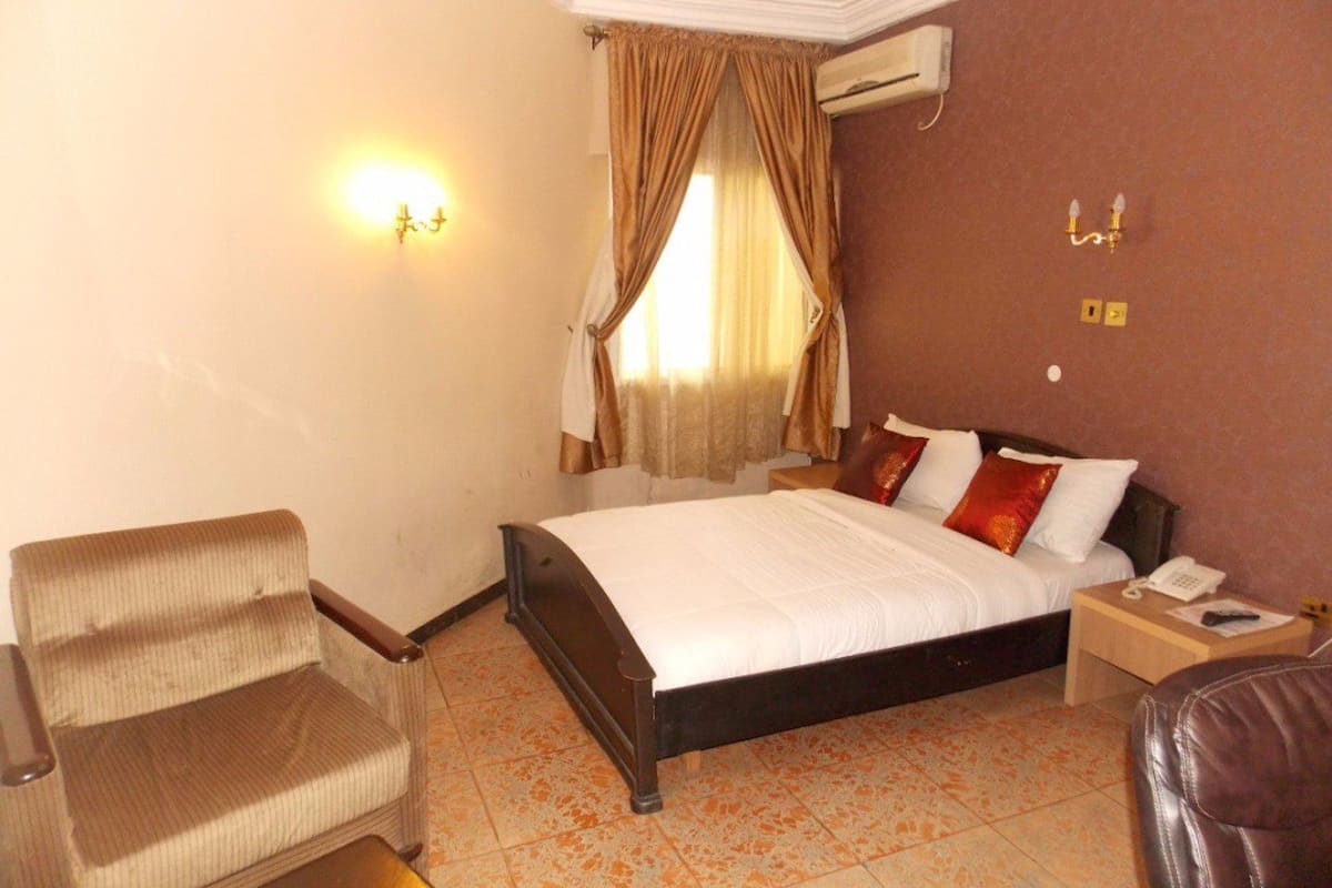 A hotel room with a bed, a chair, a nightstand, and a wall-mounted air conditioner.