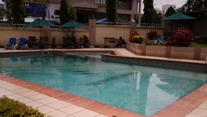 Una piscina con bordes de azulejos, rodeada de sillas de descanso y sombrillas, y un edificio al fondo.