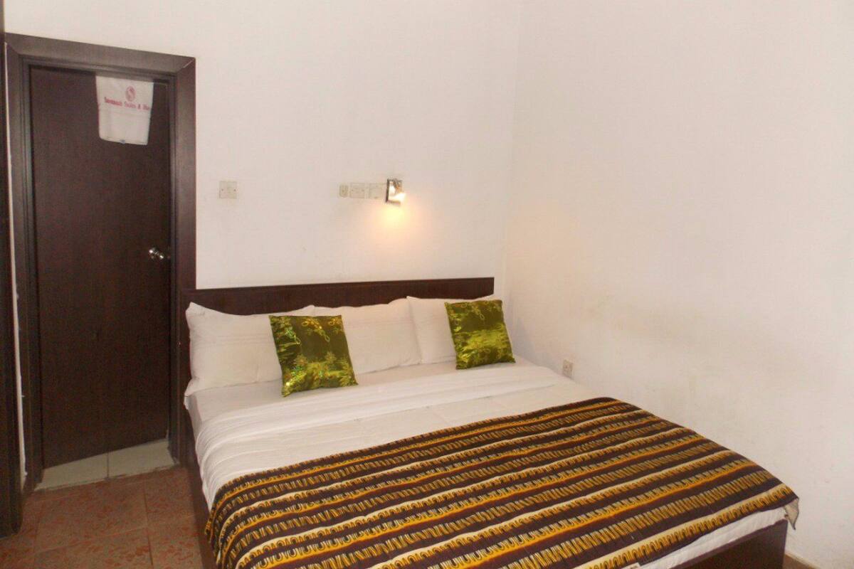 A bedroom with a bed, a nightstand, a wall-mounted light, and a door with a sign.