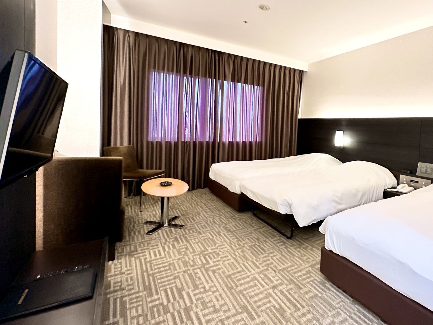 A hotel room with a bed, a desk, a chair, a TV, and a window with curtains.