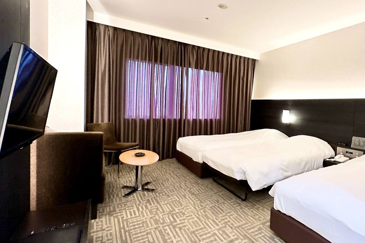 A hotel room with a bed, a desk, a chair, a TV, and a window with curtains.
