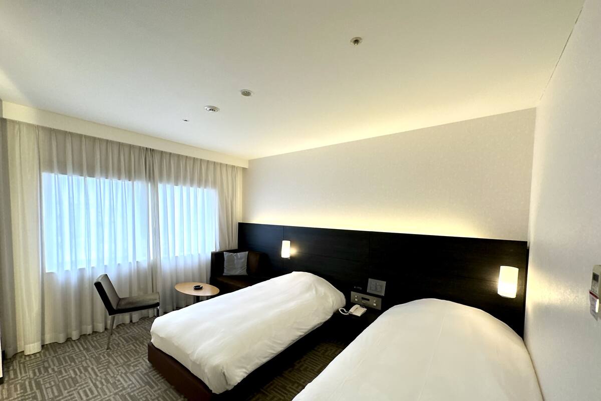 Une chambre d’hôtel avec deux lits simples, un bureau avec une chaise, une petite table et une fenêtre avec des rideaux.
