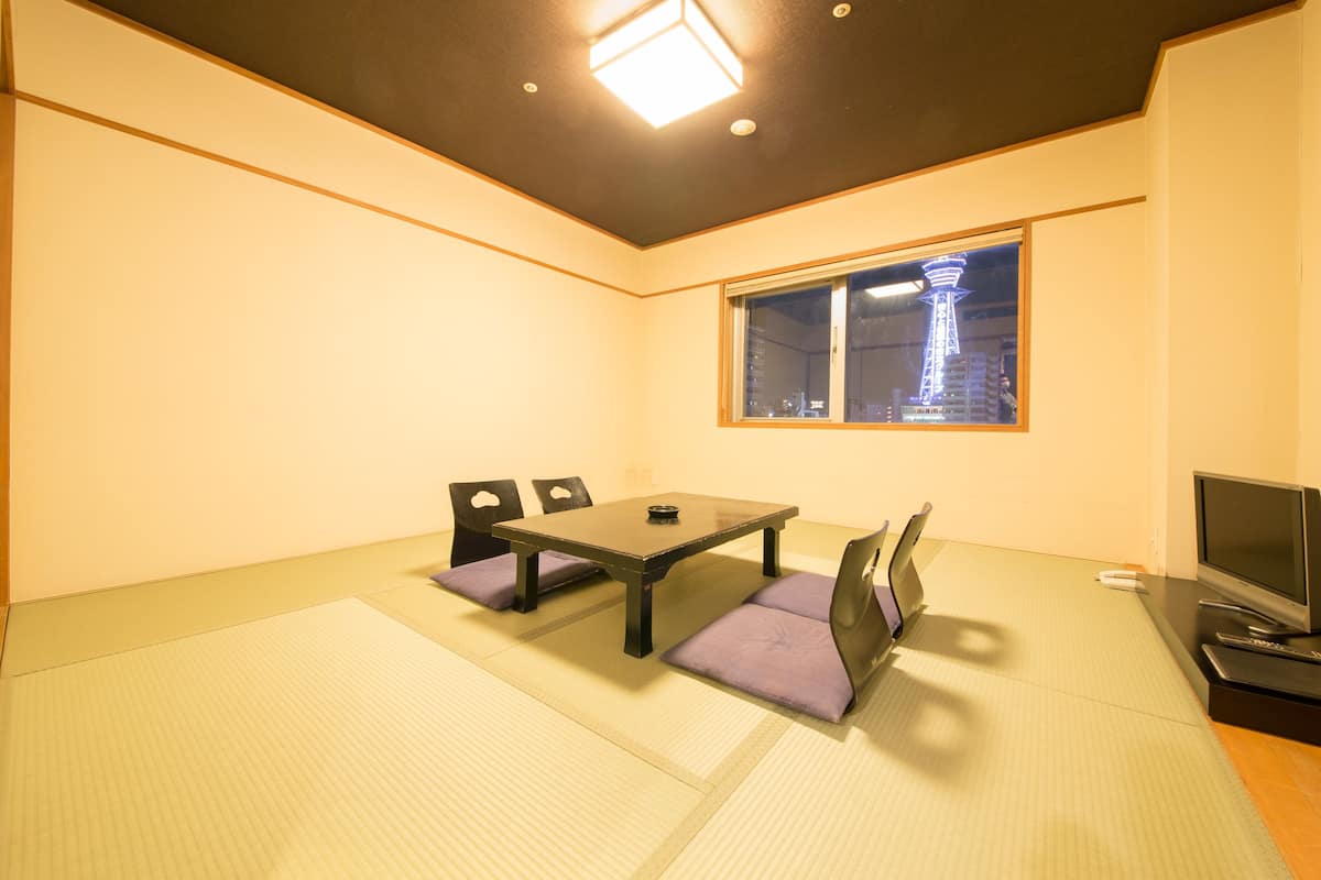 Une pièce avec un sol recouvert de tatamis, une table basse avec des coussins, un téléviseur et une fenêtre donnant sur une tour.