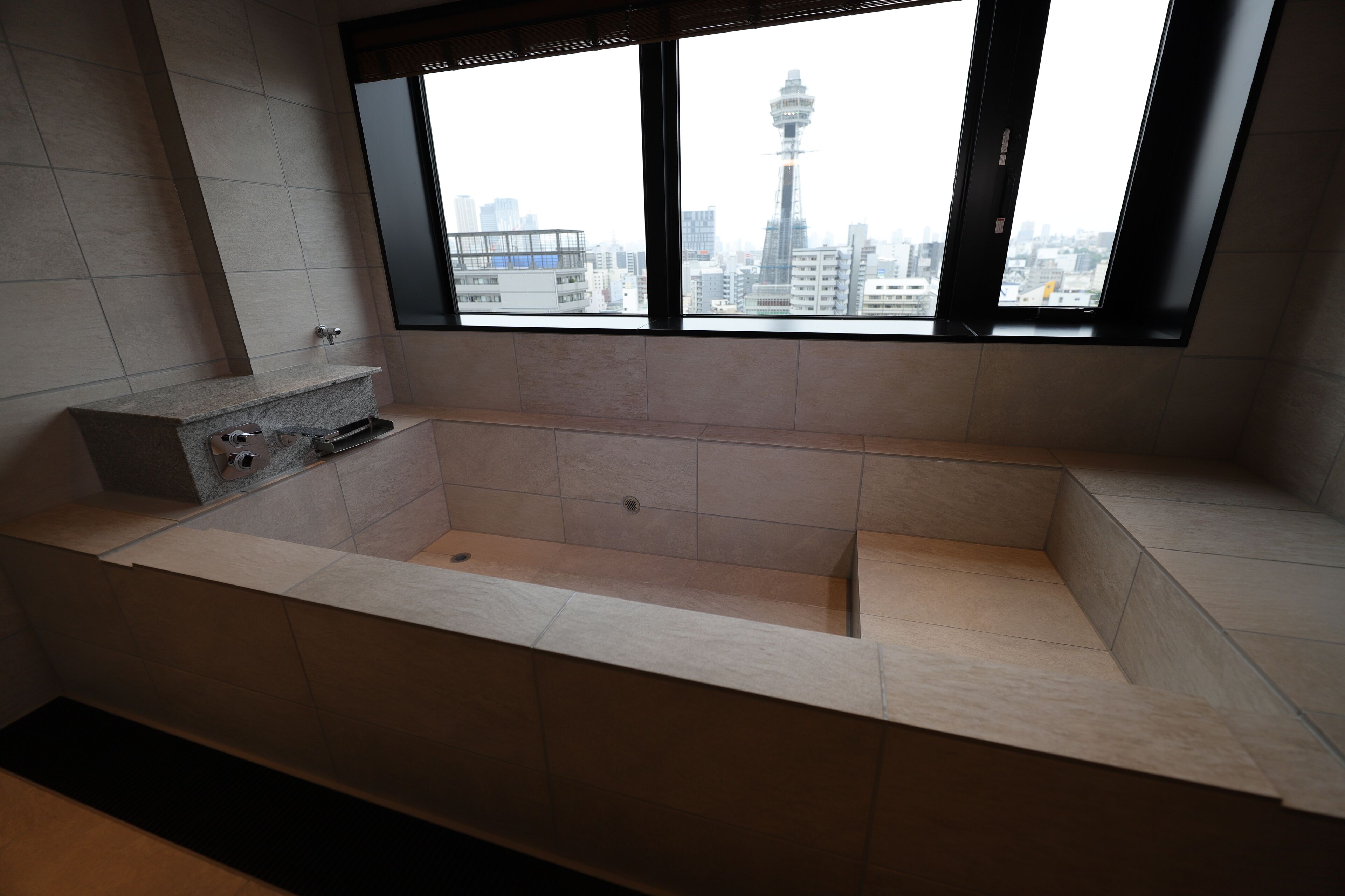 A modern bathroom with a large bathtub, a window with a city view, and a tiled floor.