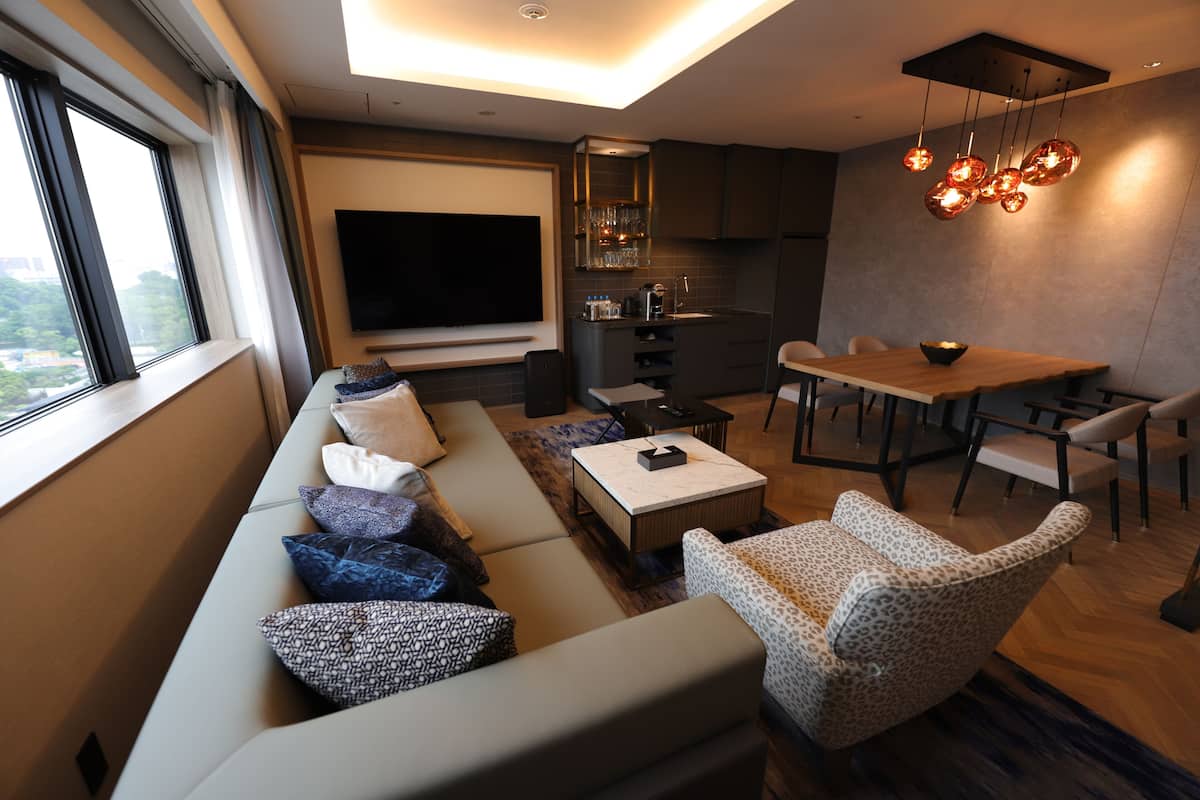 Un salon moderne avec un canapé, une table basse et des coussins à motifs. Un coin repas avec une table et des chaises est situé à côté. Un téléviseur à écran plat est fixé au mur, et on aperçoit un coin cuisine à l’arrière-plan.
