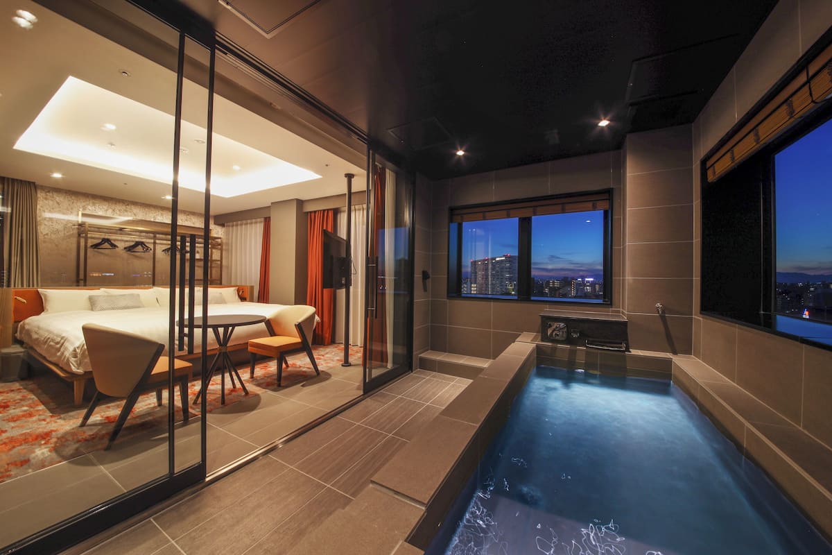 Une chambre d’hôtel moderne dotée d’une salle de bain entièrement vitrée, d’un balcon privé offrant une vue sur le paysage urbain et d’un petit espace piscine.