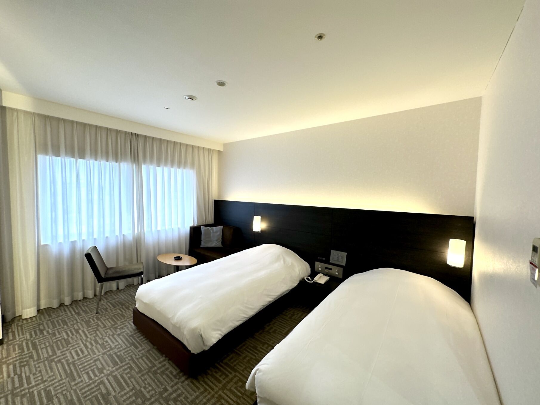 A hotel room with two beds, a desk, a chair, a lamp, and a window with curtains.