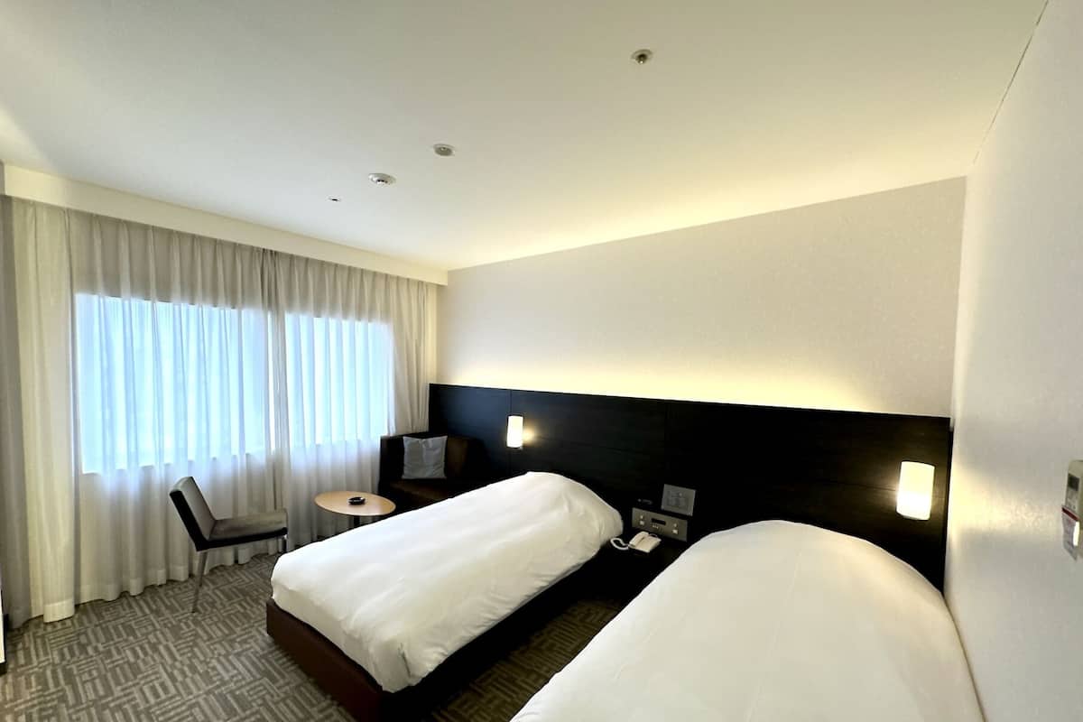 Une chambre d’hôtel avec deux lits, un bureau, une chaise, une lampe et une fenêtre avec des rideaux.