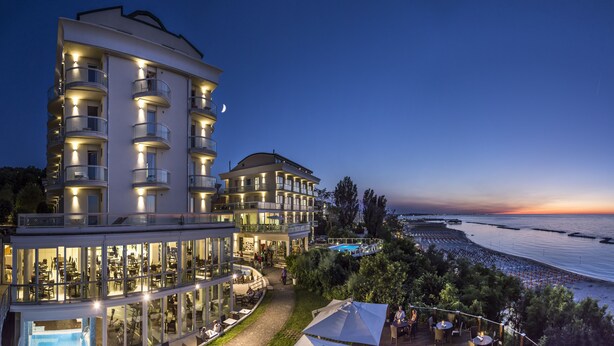 A multi-story hotel with outdoor dining areas, a pool, and a view of the sea at dusk. - Hotel Sans Souci (Gabicce Mare)