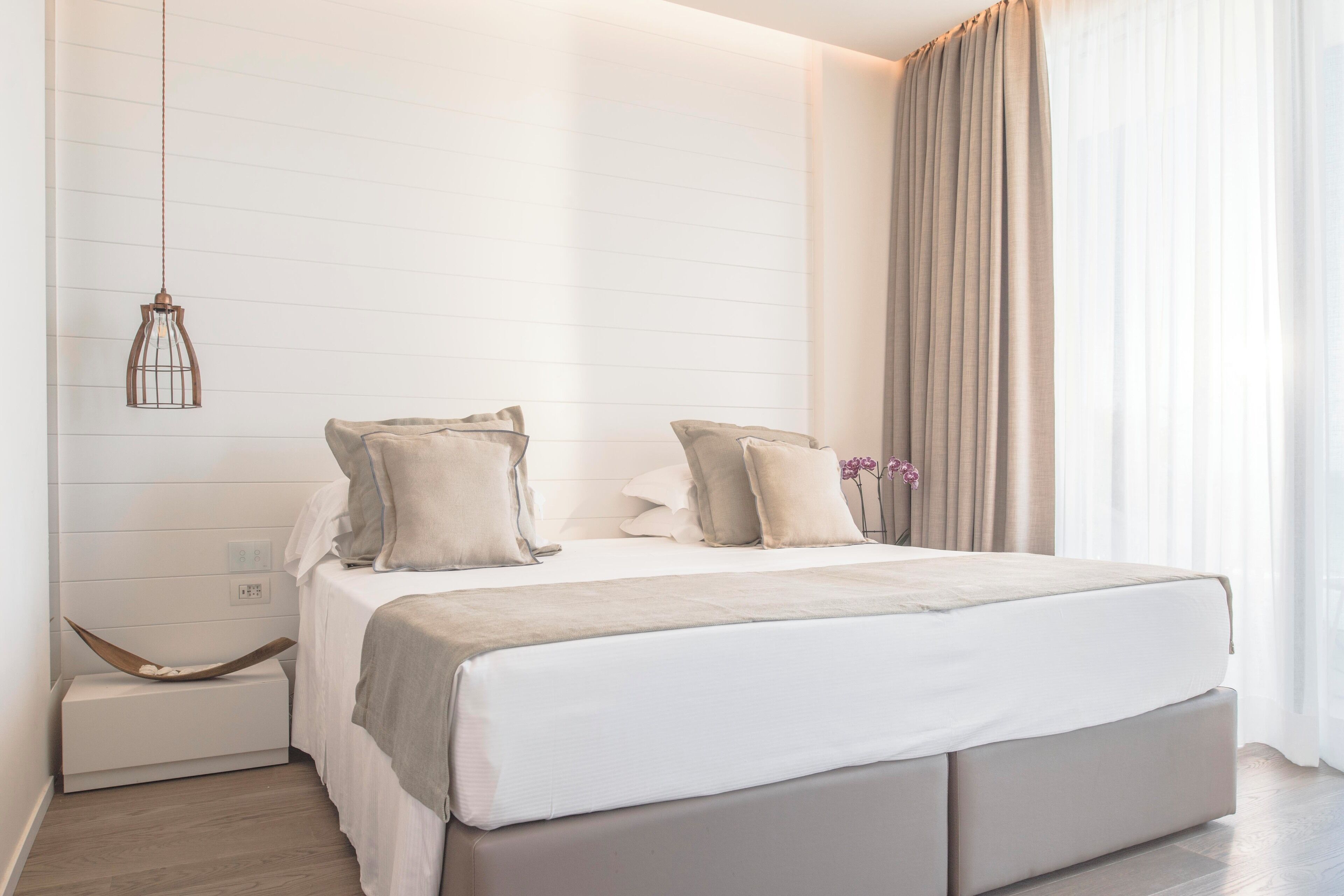 A modern bedroom with a large bed, a hanging light fixture, and a view of the outdoors through sheer curtains.