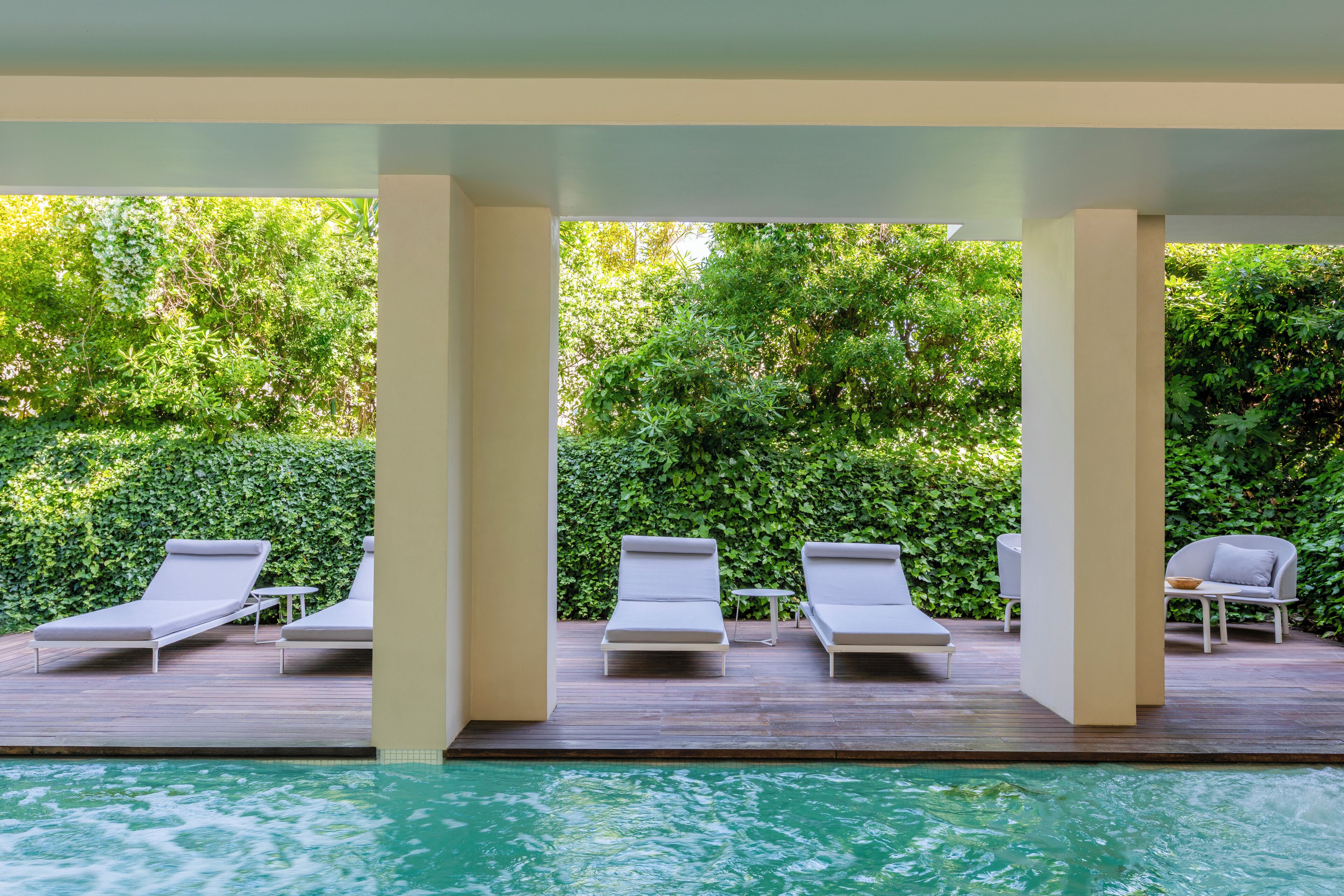 Zona junto a la piscina con sillones de descanso y una mesa, rodeada de abundante vegetación.