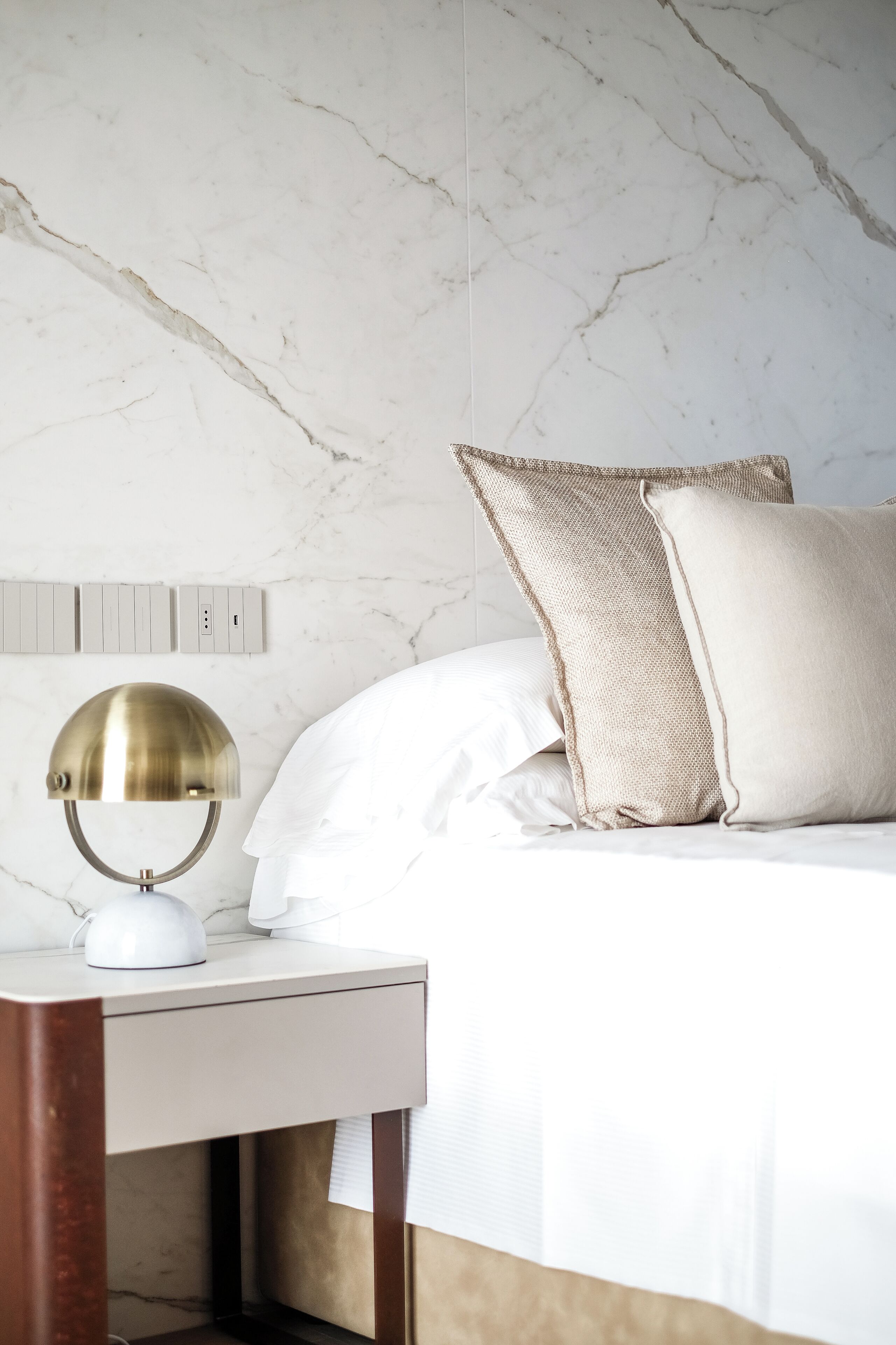 Una cama bien hecha con ropa blanca, una lámpara de escritorio de latón y una mesita de noche de madera.