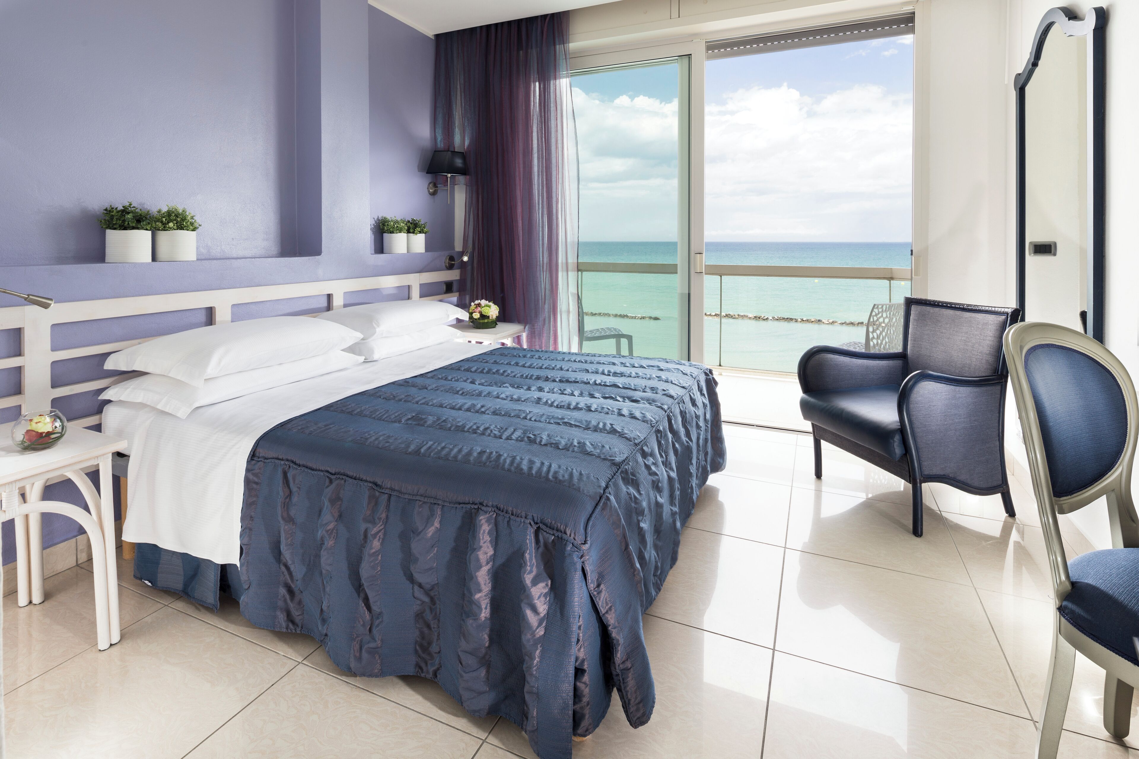 A modern hotel room with a large bed, a chair, a small table, and a balcony with a view of the sea.