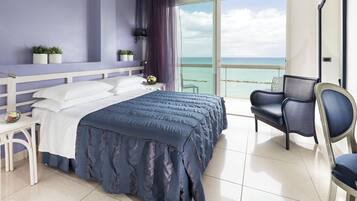 A modern hotel room with a large bed, a chair, a small table, and a balcony with a view of the sea.