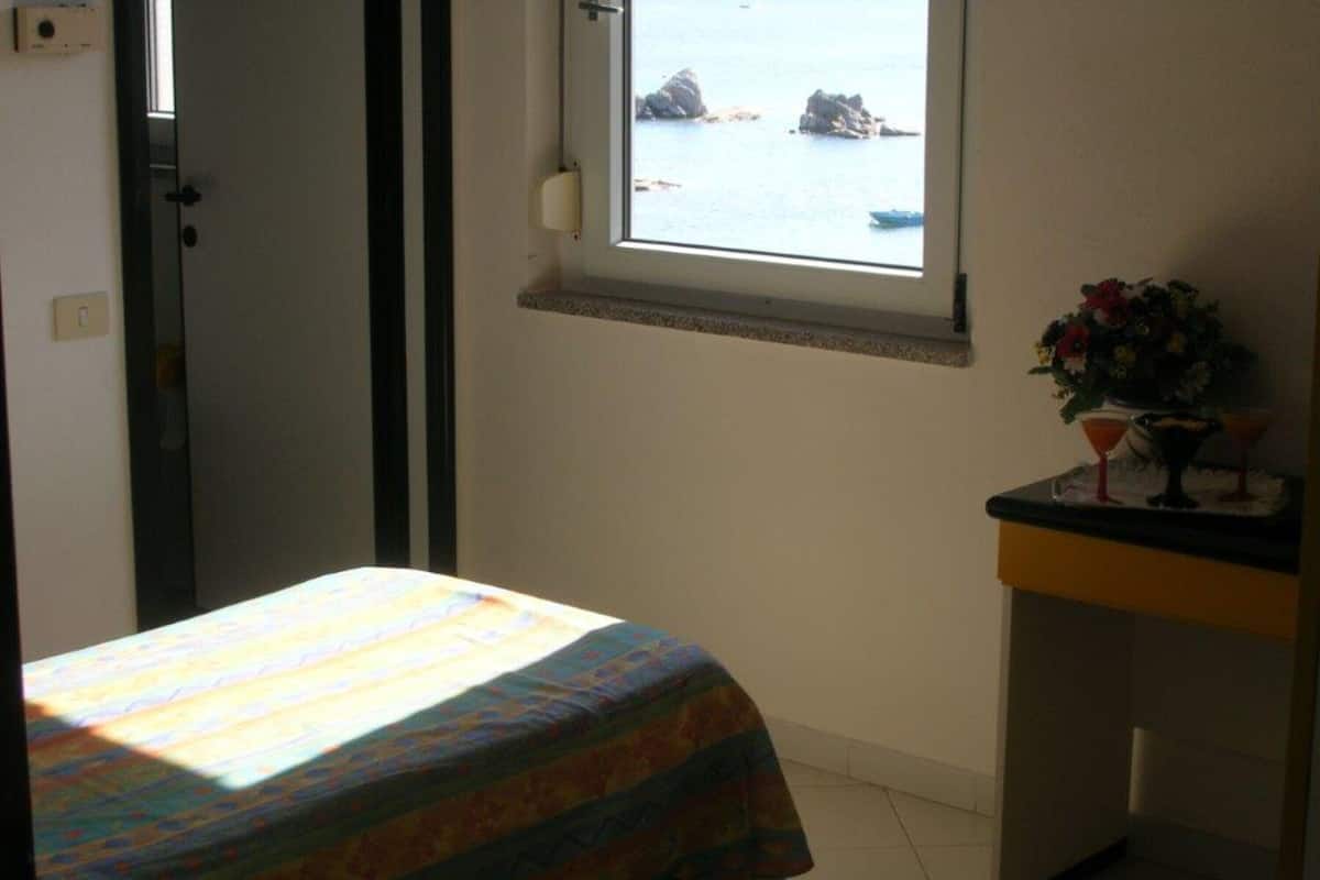 A room with a bed, a small desk with a vase of flowers, and a window overlooking a body of water.
