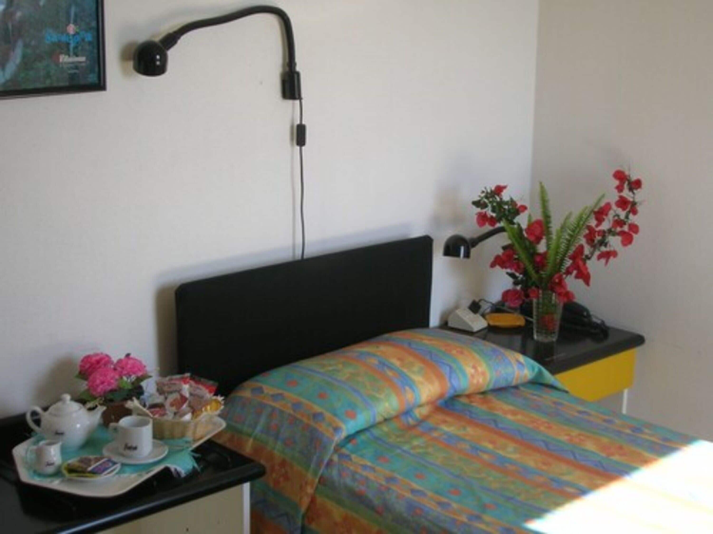 A room with a bed, a desk with a lamp, a vase with flowers, and a framed picture on the wall.
